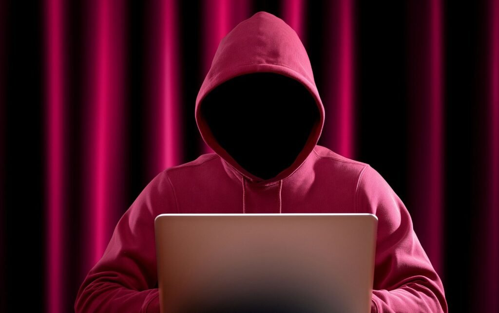Un pirate informatique cagoulé en sweat mauve, assis de face devant un ordinateur.